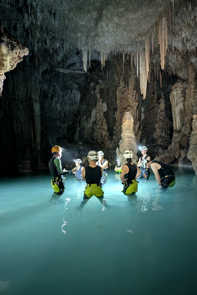 Teilnehmer der Cova des Coloms Tour stehen im klaren Wasser der Höhle mit beleuchteten Helmen und Tropfsteinen