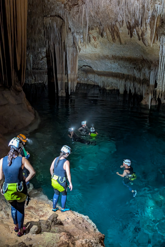 Teilnehmer der Cova des Coloms Tour schwimmen durch das türkisfarbene Wasser der Höhle mit Tropfsteinformationen